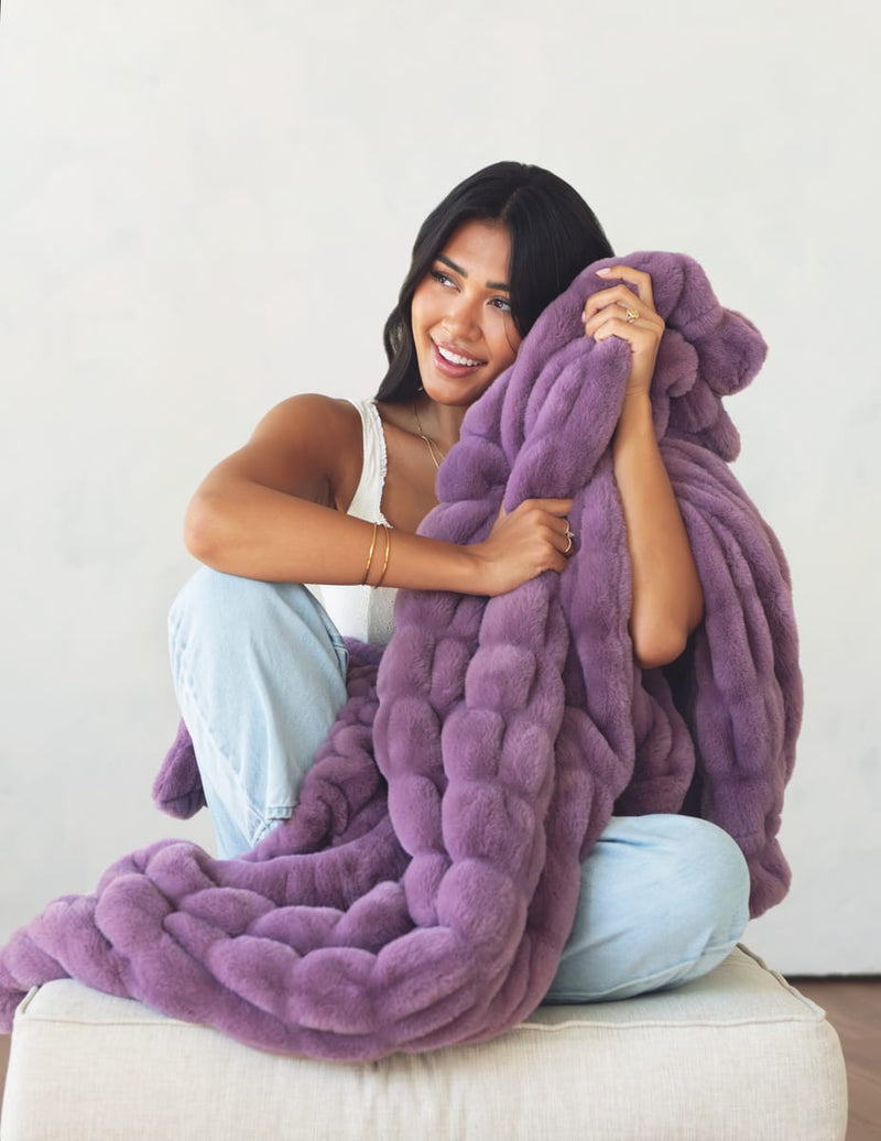 A woman sitting on a white ottoman holds a chunky, oversized plush throw blanket in a dusty purple color against a plain off-white wall.
