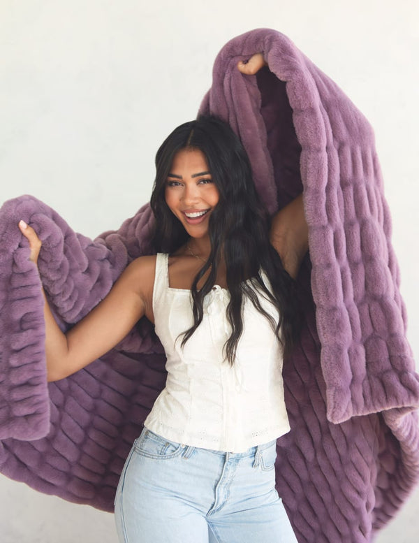 A woman with long dark hair smiles while holding up a chunky, ribbed plush throw blanket in a dusty purple color against a plain off-white background.