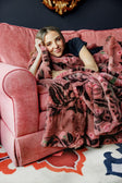 A woman reclining on a pink velvet sofa, wrapped in a plush pink floral throw blanket with black and green leaf patterns. The setting includes a dark navy wall, a gold decorative mirror, and a patterned rug.