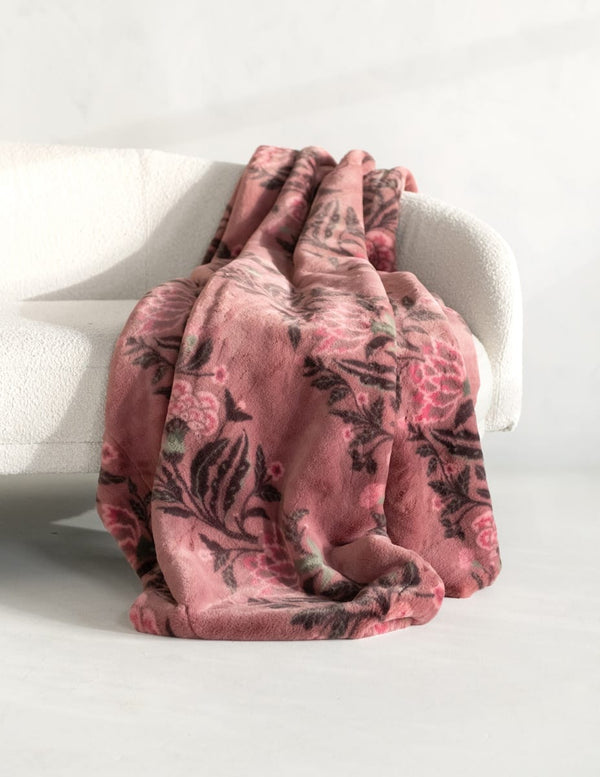 A plush throw blanket with a dusty pink and black botanical floral pattern draped over a white armchair in a bright, minimalist room.