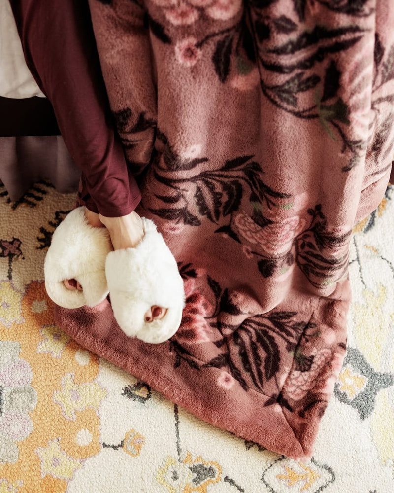 A cozy close-up of a dusty rose floral  blanket draped over a person's legs, with fluffy white slippers on a patterned rug.