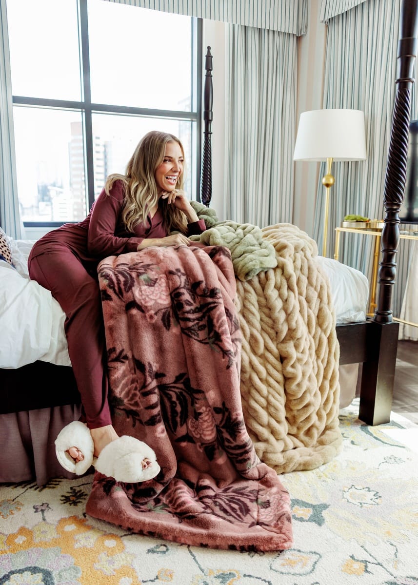 A woman lounges on a bed in a bright bedroom, wrapped in a dusty rose floral plush blanket with a beige plush knit throw and white slippers. She wears matching burgundy pajamas, and the room has striped curtains, a window view, and a floor lamp.