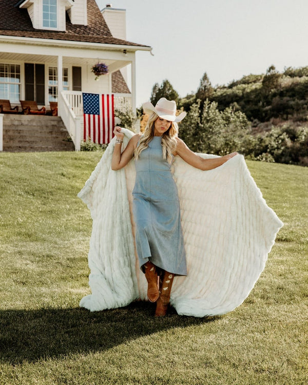 A woman in a light blue dress and cowboy hat stands on a grassy lawn holding up a large white textured blanket, with a white house, American flag, and hills in the background.