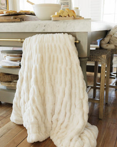 A chunky throw blanket in off-white is draped over the leg of a kitchen island table, with a marble countertop, wooden stools, and a warm wood floor visible in the background.