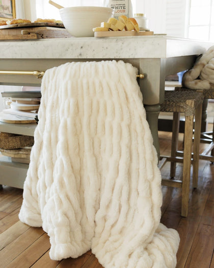A chunky throw blanket in off-white is draped over the leg of a kitchen island table, with a marble countertop, wooden stools, and a warm wood floor visible in the background.