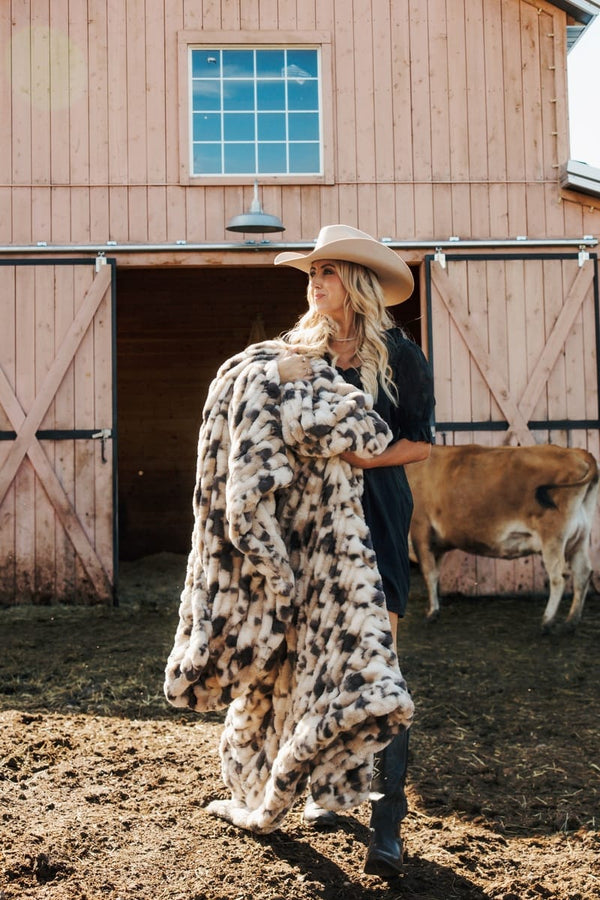 A woman in a cowboy hat and dark dress walks in front of a barn holding a large faux-fur blanket with a black, white, and beige pig-print pattern. 