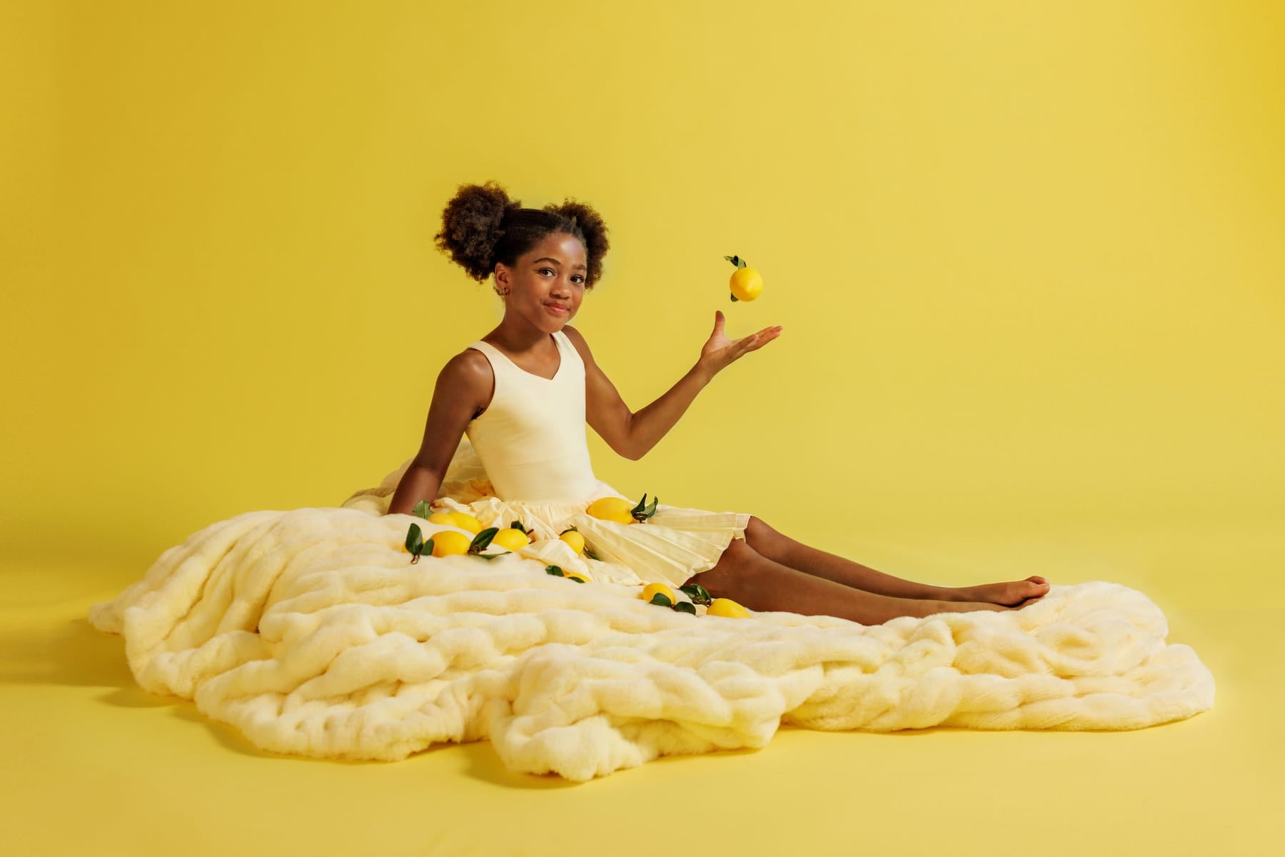 Young girl sitting on a yellow blanket while holding lemons against a yellow background.