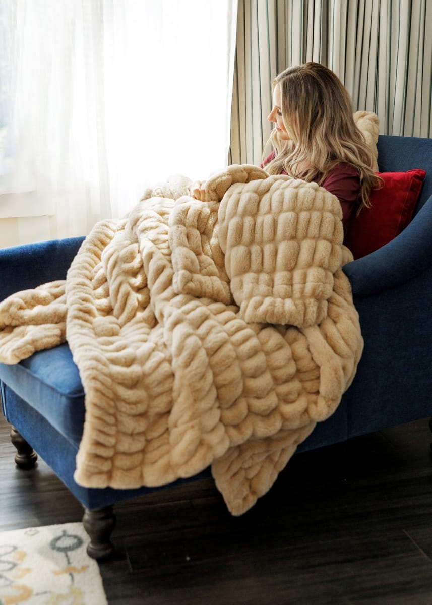 A woman sits on a blue armchair in a living room, wrapped in a chunky plush beige throw blanket. The blanket has a thick, textured boucle-like pattern and is draped over the chair and floor.