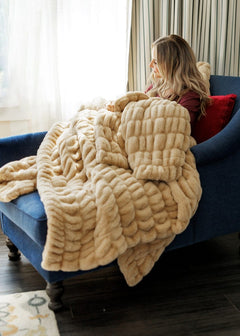 A woman sits on a blue armchair in a living room, wrapped in a chunky plush beige throw blanket. The blanket has a thick, textured boucle-like pattern and is draped over the chair and floor.