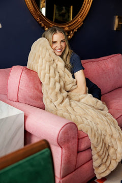 A woman smiling while wrapped in a chunky knit beige throw blanket, sitting on a pink sofa in a styled living room.