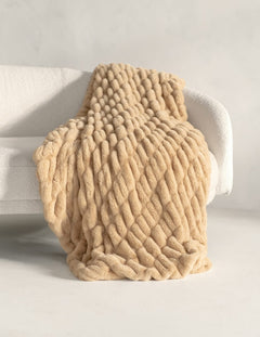A chunky, thick-knit beige throw blanket draped over a white armchair in a bright, minimalist room.