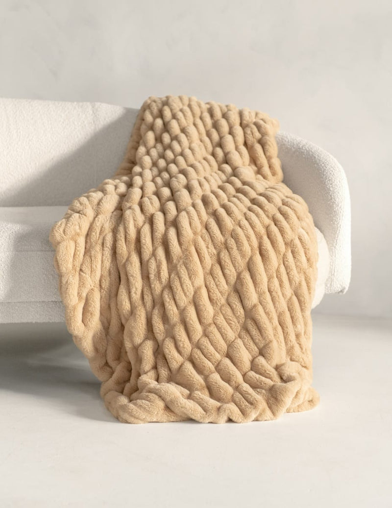A chunky, thick-knit beige throw blanket draped over a white armchair in a bright, minimalist room.
