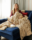A woman sits on a blue armchair wrapped in a chunky, thick tan-colored knit blanket.