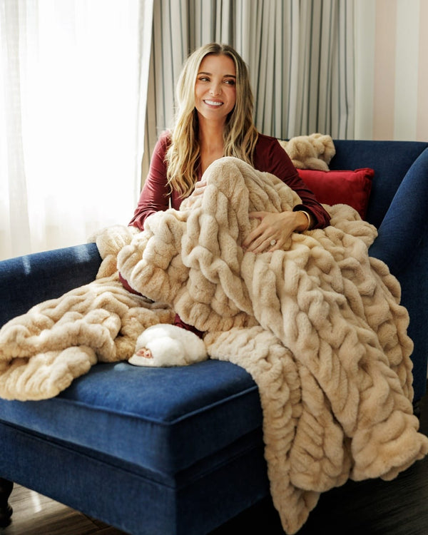 A woman sits on a blue armchair wrapped in a chunky, thick tan-colored knit blanket.