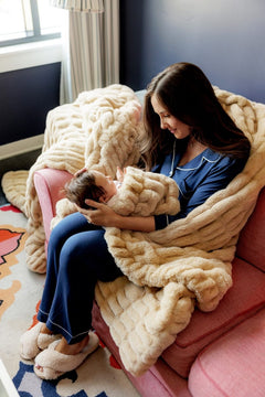 A woman in blue pajamas sits on a pink couch, holding and swaddling a baby in a chunky tan knit blanket. A matching oversized chunky knit throw blanket is draped over the couch in a cozy living room setting.