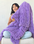 A woman sitting on a light-colored sofa wrapped in a chunky, plush purple blanket. The blanket has a thick, ribbed texture and is draped over her and the sofa against a plain off-white wall.