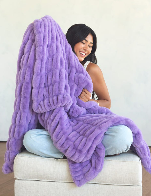 A woman sitting on a light beige ottoman wrapped in a chunky, oversized lavender purple throw blanket. The blanket has a thick, ribbed texture and is draped over her and around the ottoman against a plain off-white wall.