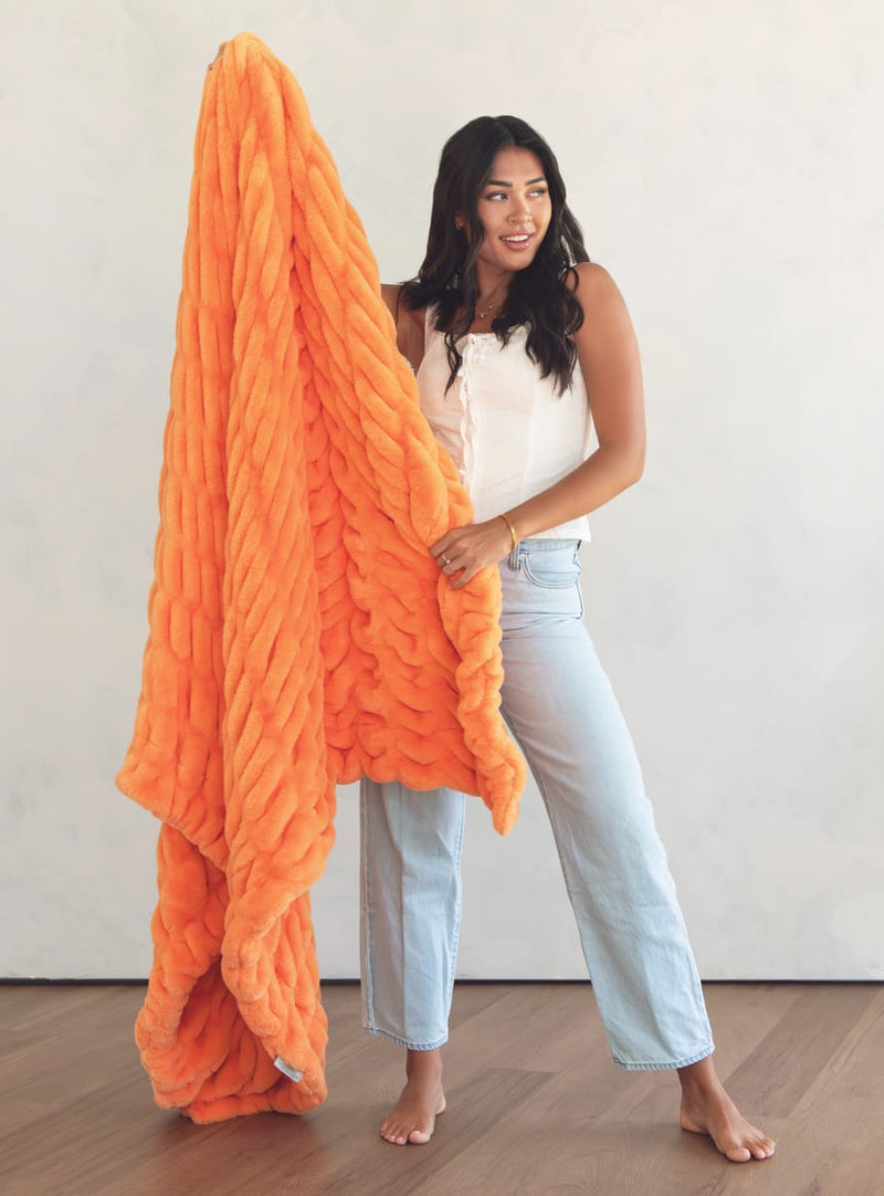 A woman stands barefoot in a room with a light wall and wood floor, holding up a orange throw blanket. She is wearing a white sleeveless top and light blue jeans.