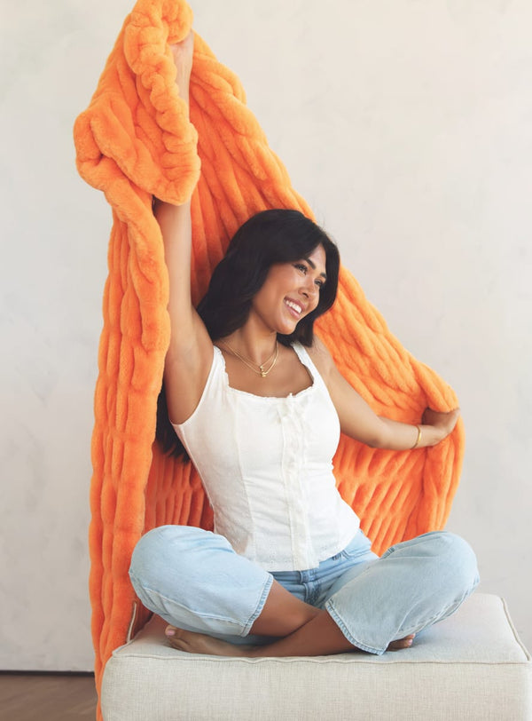A woman sitting cross-legged on a light beige upholstered bench, smiling while holding up a chunky, plush orange blanket against a plain off-white wall.