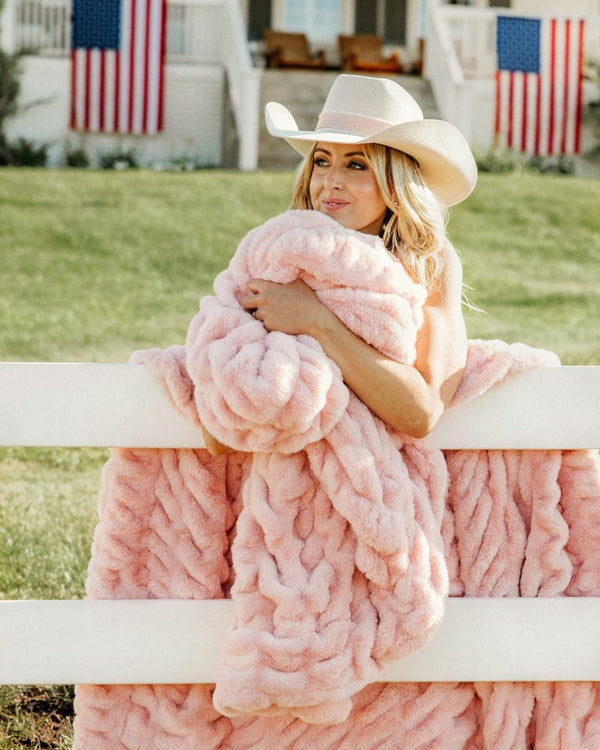 A woman in a white cowboy hat sits on a white fence outdoors, wrapped in a chunky, plush pink blanket. American flags and a white house are visible in the background.