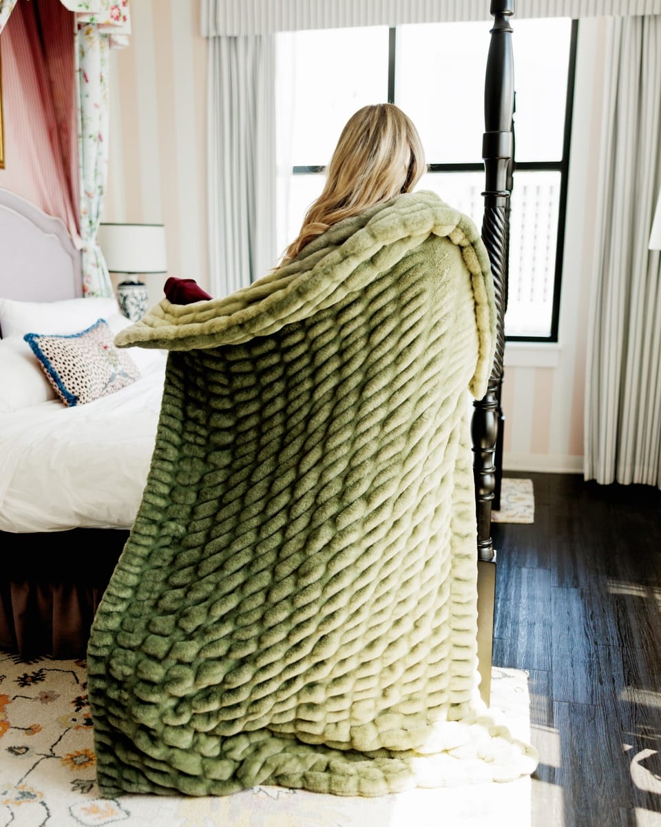 A woman stands with her back to the camera, wrapped in a chunky, ribbed sage green ombre throw blanket in a bright bedroom with a bed, window, and dark wood floor.