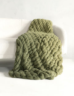 A chunky, thick-knit ombre olive green throw blanket draped over a white armchair in a bright, minimalist room.