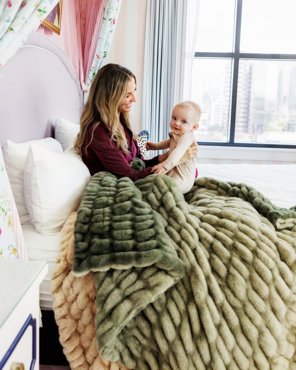 A woman and a baby sit together on a bed in a bright bedroom, covered by a chunky knit throw blanket in sage green and beige. The room has a large window with a city view, white pillows, and floral curtains.