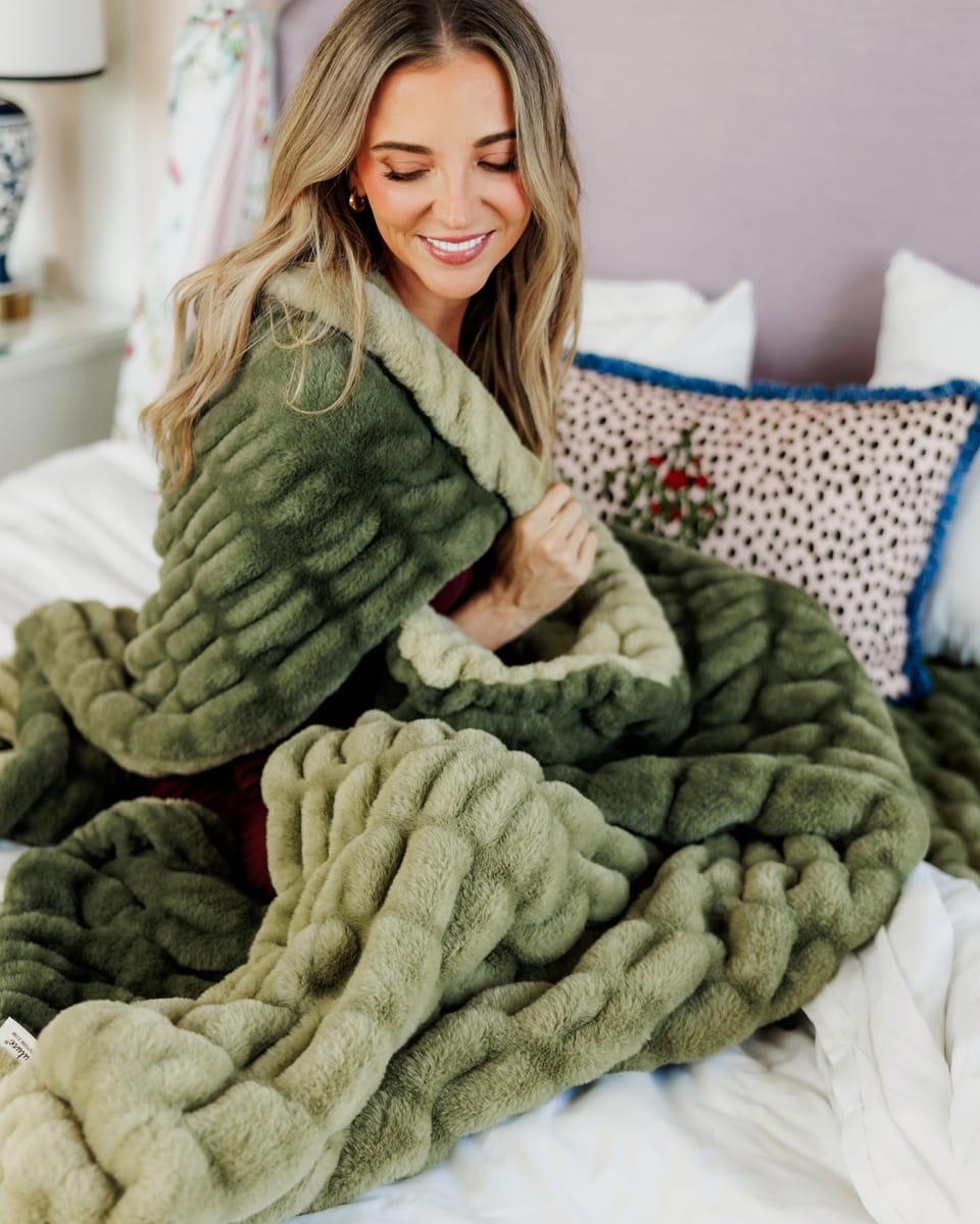 A woman sitting on a bed wrapped in a chunky, ribbed plush throw blanket. The blanket is olive green with an ombre effect.