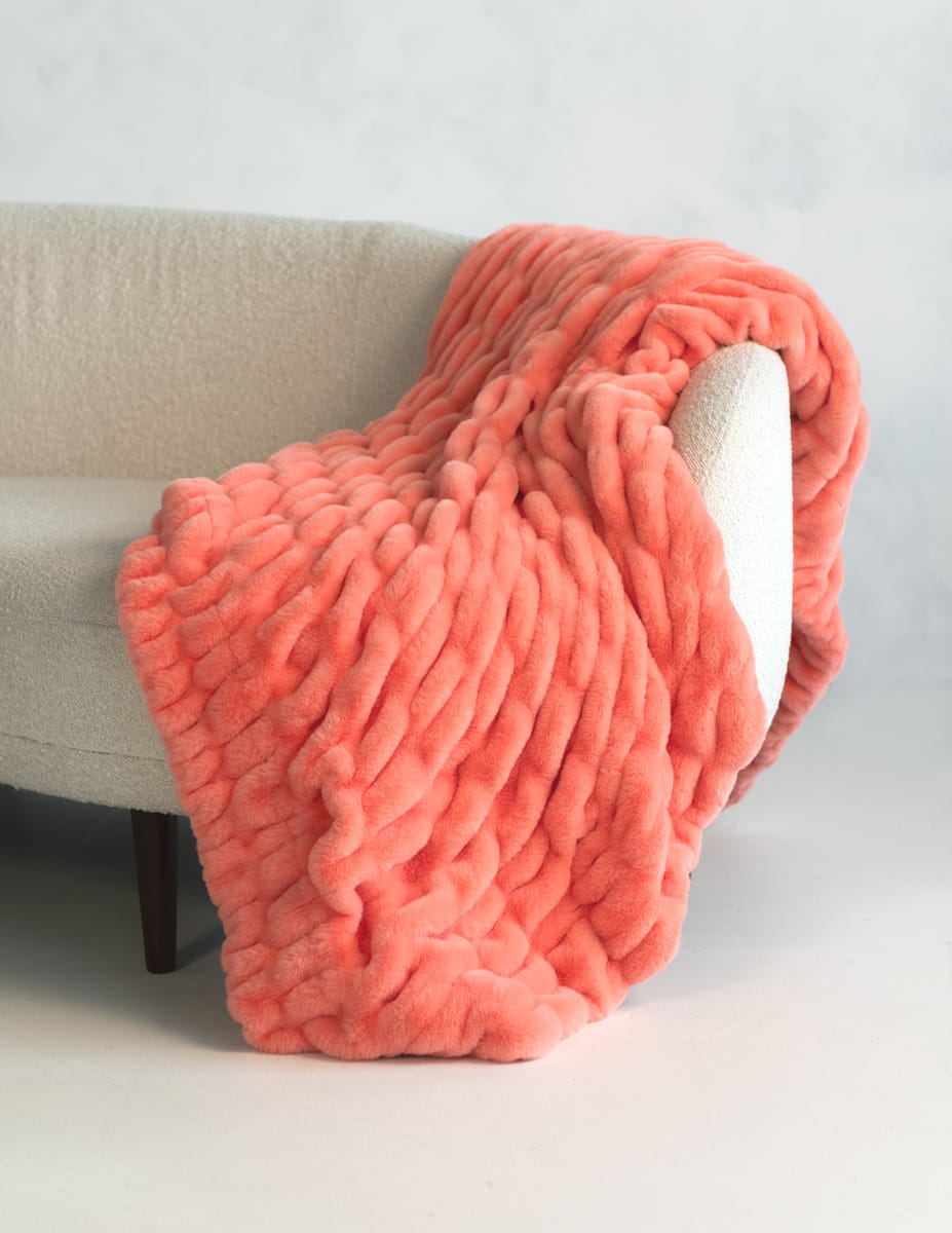 A chunky, thick-textured throw blanket in a coral pink color, draped over the arm of a light beige upholstered armchair.