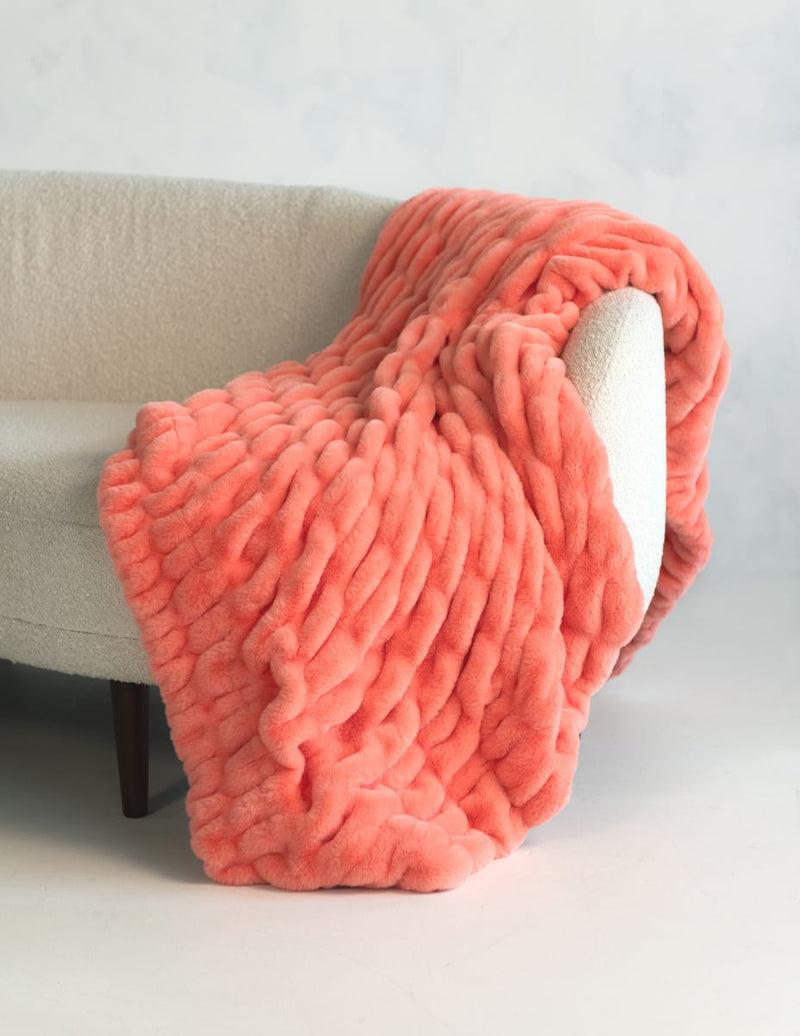 A chunky, thick-textured throw blanket in a coral pink color, draped over the arm of a light beige upholstered armchair.