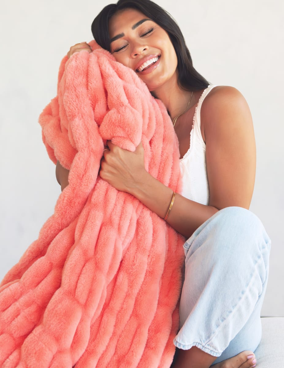 A woman smiling while hugging a chunky, oversized coral pink faux fur blanket.