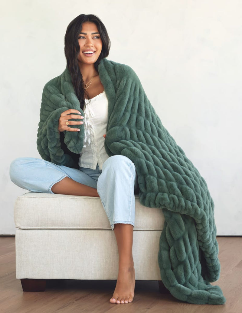 A woman sits barefoot on a cream upholstered ottoman, wrapped in a  sage green blanket. 