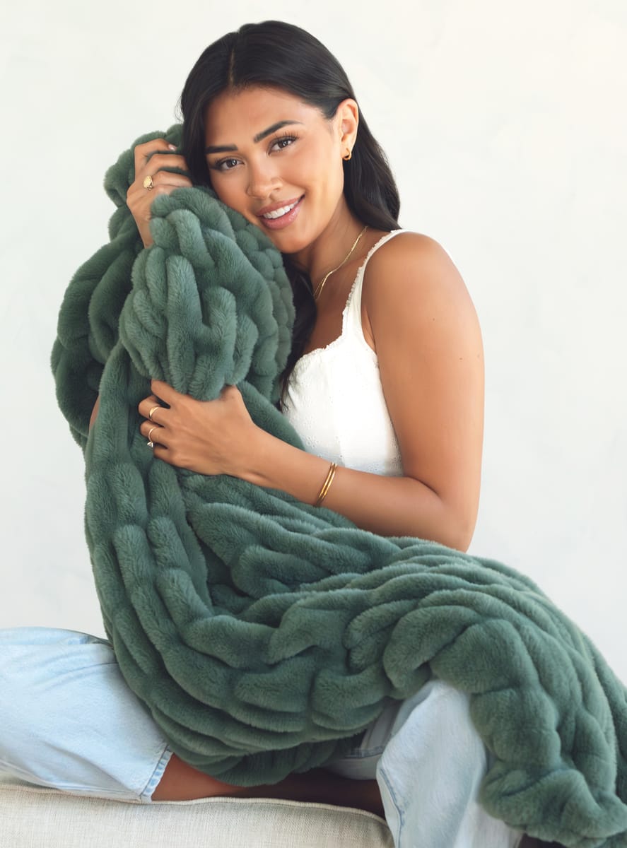 A woman sitting on a light-colored sofa, holding a throw blanket in a dusty sage green color.