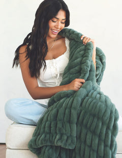 A woman with long dark hair sits on a white upholstered bench, smiling while holding up a chunky, oversized plush throw blanket in a muted sage green color.