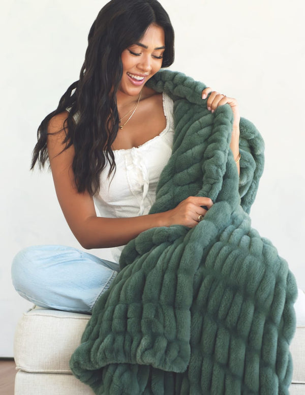 A woman with long dark hair sits on a white upholstered bench, smiling while holding up a chunky, oversized plush throw blanket in a muted sage green color.