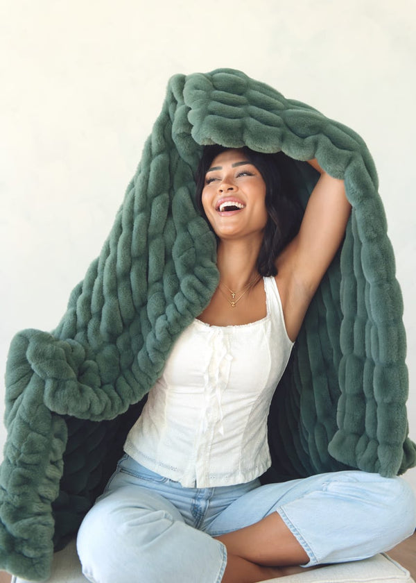 A woman sitting cross-legged under a chunky, oversized dark green plush blanket, wearing a white top and light blue jeans against a plain off-white background.