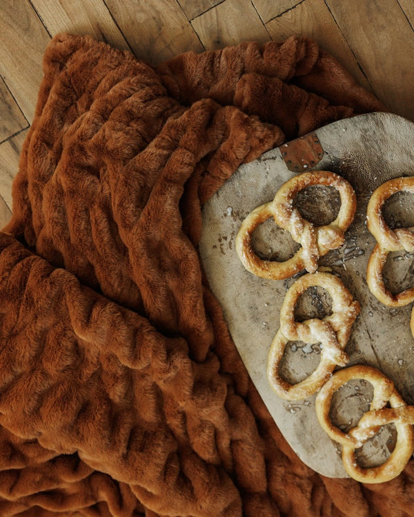 A close-up of a chunky, textured brown throw blanket draped over a rustic stone plate holding several pretzels, set on a wooden floor.