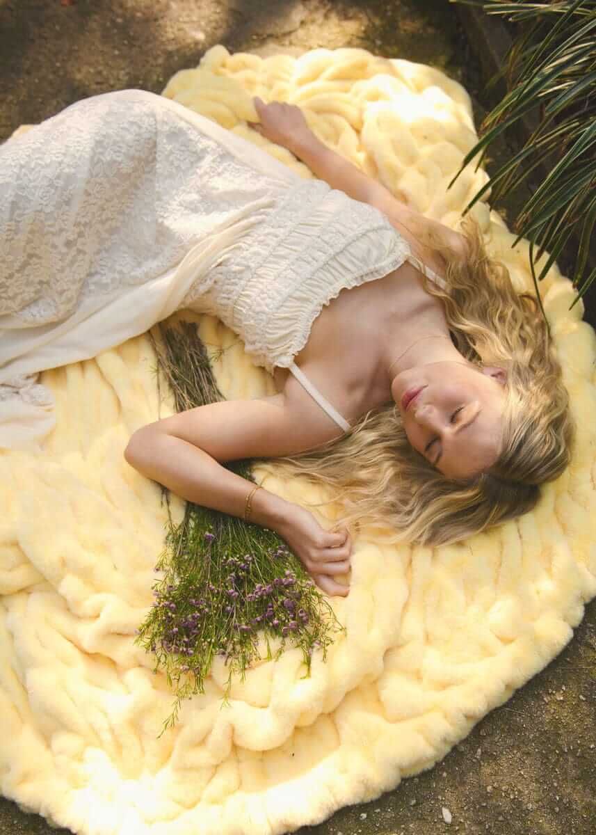 A woman lying on a Hugs Butter Yellow Blanket, surrounded by lavender flowers, enjoying a soft and cozy moment outdoors.