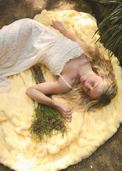 A woman lying on a Hugs Butter Yellow Blanket, surrounded by lavender flowers, enjoying a soft and cozy moment outdoors.