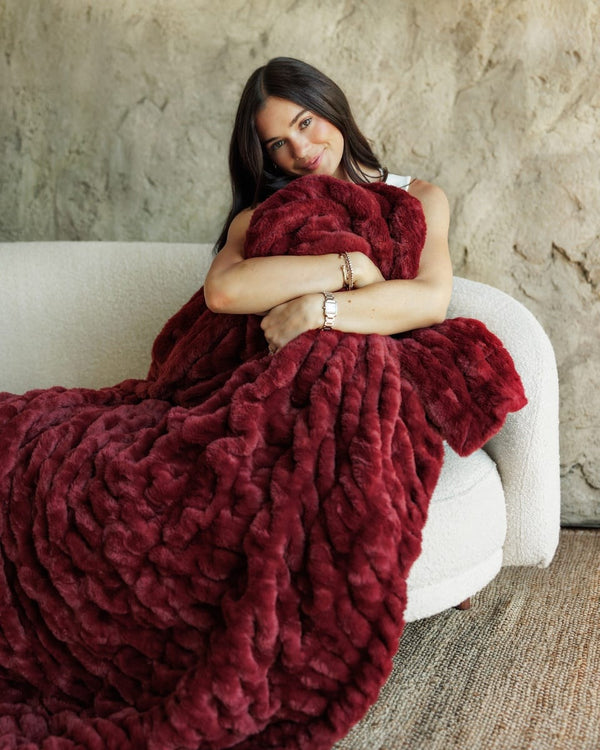 A woman sits on a white textured sofa wrapped in a chunky, Hugs Cabernet Blanket, set against a rustic beige stone wall.