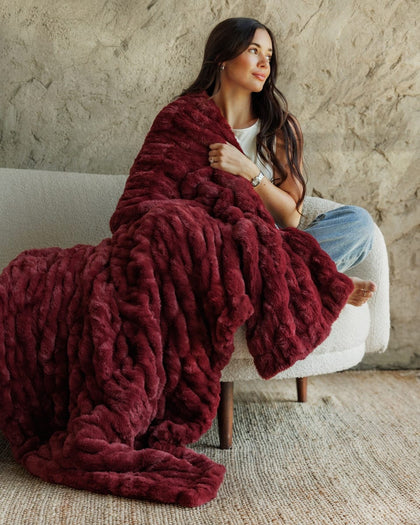 A woman sits on a white armchair wrapped in a chunky, Hugs Cabernet Blanket, set against a textured concrete wall and woven rug.