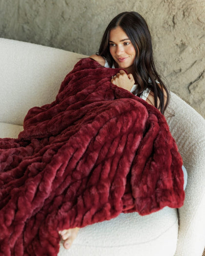 A woman sits on a cream-colored sofa, wrapped in a chunky, Hugs Cabernet Blanket. The background features a textured, warm gray wall.