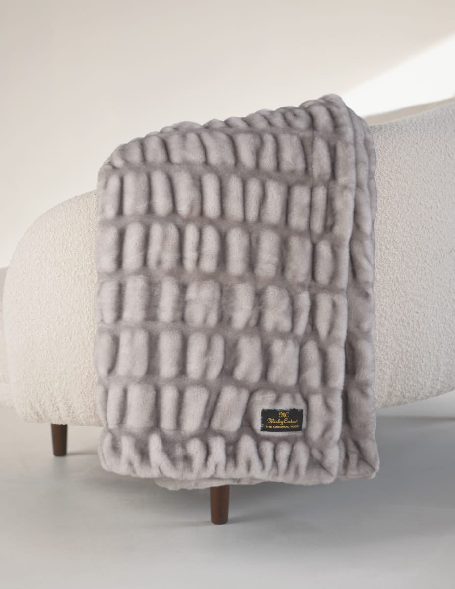 Hugs Chai Blanket draped on a cozy chair, showcasing its soft gray texture and stylish design.