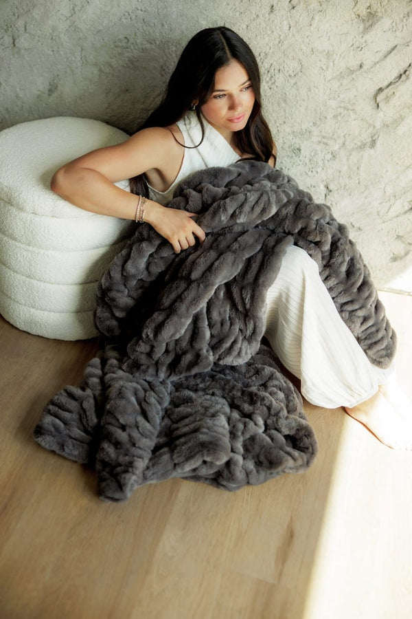 A woman reclines on a light wood floor against a textured stone wall, wrapped in a chunky, Hugs Charcoal Blanket. She is wearing a white sleeveless top and white pants, sitting next to a white textured pouf.