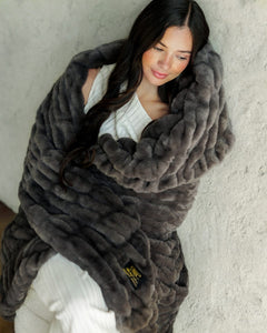 A woman wrapped in a chunky, plush Hugs Charcoal Blanket, leaning against a textured light gray wall. She is wearing a white ribbed top and white pants.
