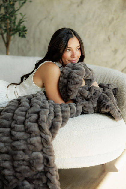 A woman reclining on a white sofa while wrapped in a Hugs Charcoal Blanket. The blanket is shown in close-up, highlighting its thick, textured boucle-like pile.