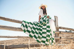 Model draped in Hugs Check Green Blanket sitting on a fence, showcasing its vibrant green and white checkered design.