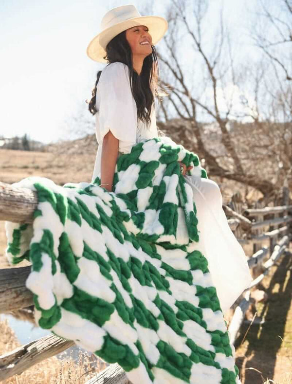 A woman wearing a white dress sits on a fence, wrapped in a Hugs Check Green Blanket with a green and white checkered pattern.