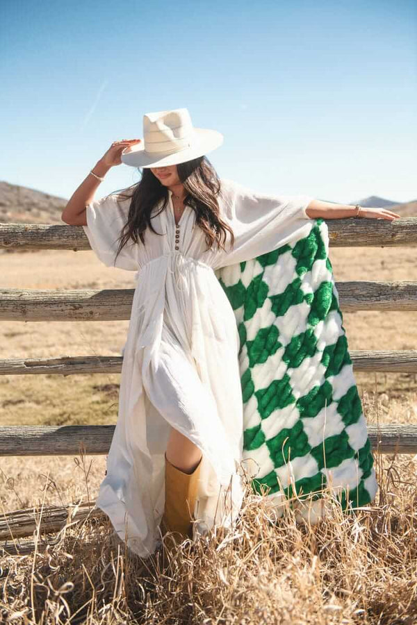 Model in a white dress holding a Hugs Check Green Blanket by a wooden fence on a sunny day.