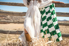 Model in white dress with Hugs Check Green Blanket draped over a fence in a rustic outdoor setting.